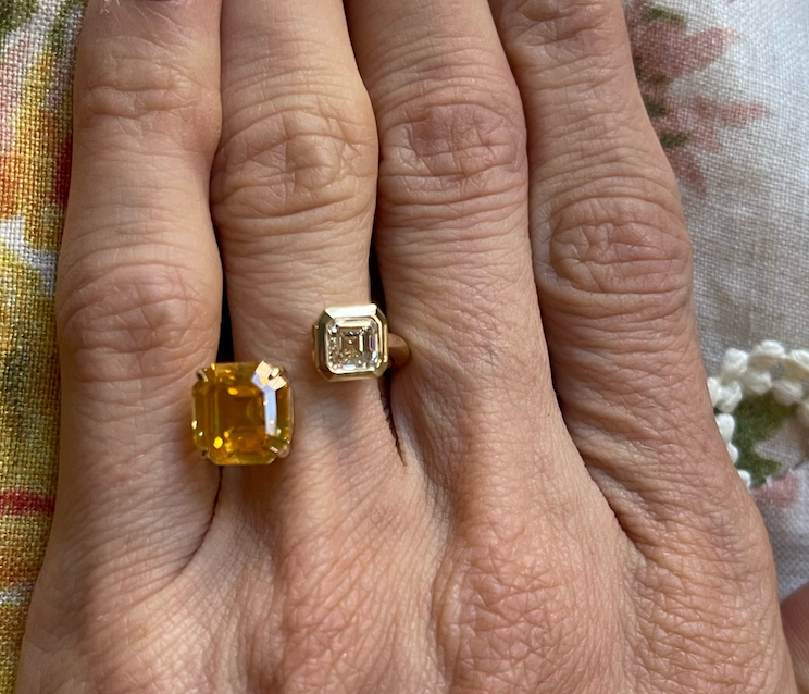A hand wearing the Yellow Sapphire and Diamond Toi et Moi Ring—featuring an 18K yellow gold yellow sapphire and a clear asscher cut diamond—rests against a floral-patterned background.