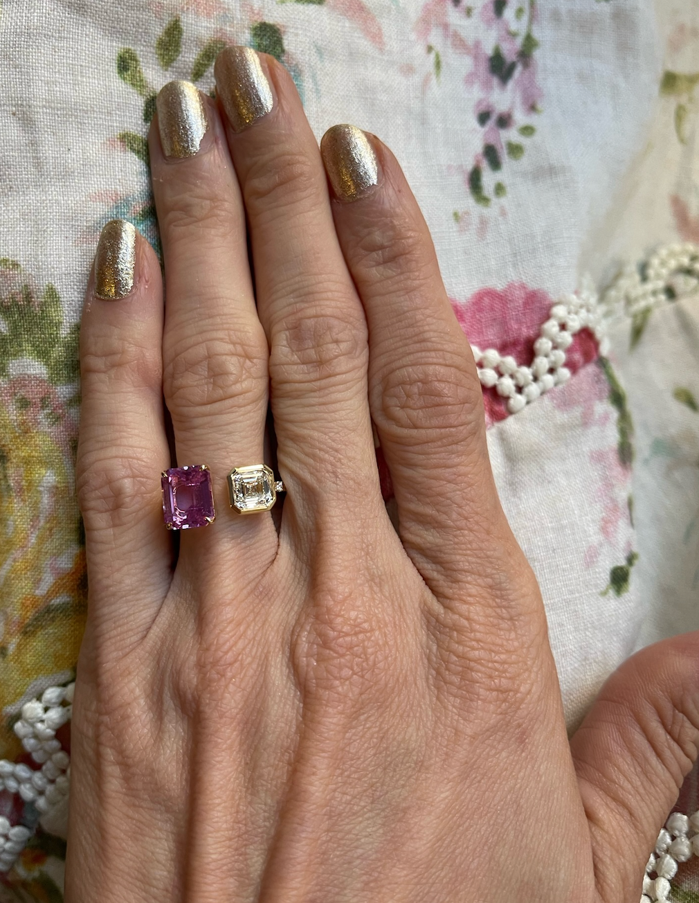 A hand with metallic gold nails wears the Toi et Moi Pavé Ring, featuring a square emerald-cut diamond and pink emerald-cut sapphire, against floral-patterned fabric with white beaded trim.