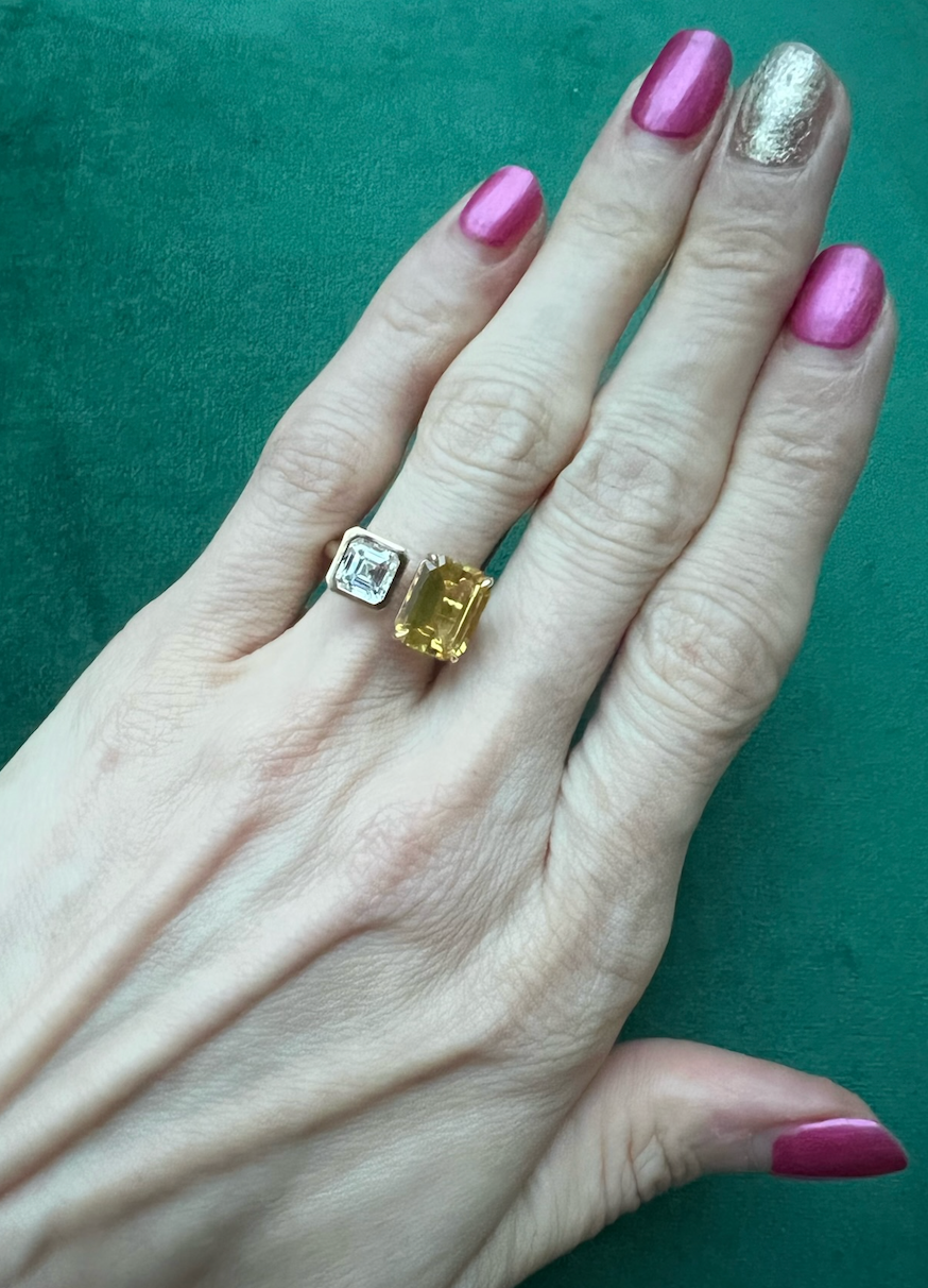 A hand with pink and gold-painted nails showcases the Toi Et Moi Ring in yellow gold, featuring a white Asscher cut diamond and a fancy yellow emerald cut diamond side by side, against a green background.