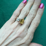 A hand with pink and gold nails wears the Toi et Moi Ring with Asscher Cut and Emerald Cut Diamonds in Yellow Gold, set against a green background.