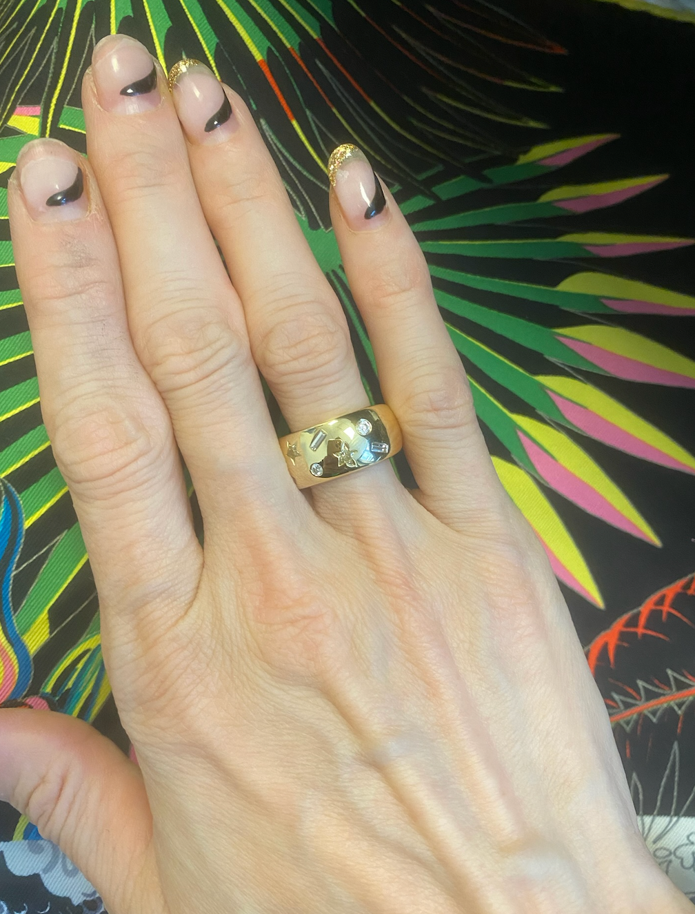 A hand wears the Yellow Diamond Celestial Cigar Band, an 18k yellow gold ring with star and gem motifs, against a colorful backdrop. The nails are manicured in black-tip French style with gold accents.