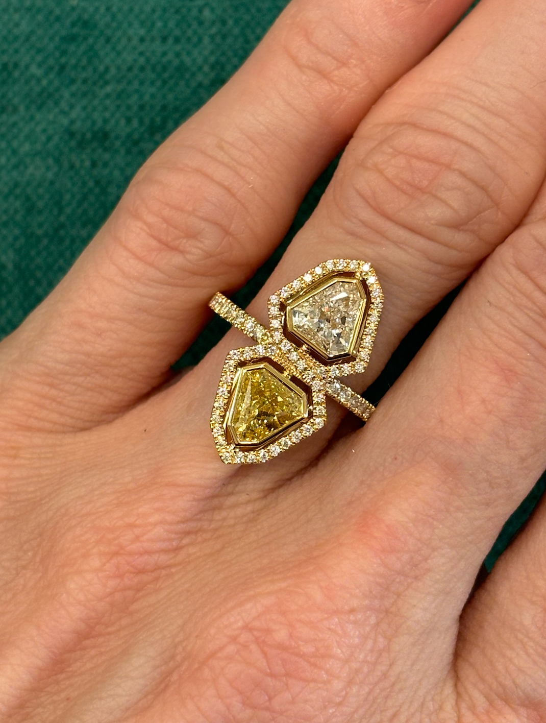 A hand displays the Yellow Diamond and White Diamond Double Shield Ring with Halo in Yellow Gold, featuring two large uniquely-shaped gems—one white diamond, one yellow diamond—each encircled by smaller diamonds, set against green fabric.