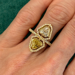 A hand displays the Yellow Diamond and White Diamond Double Shield Ring with Halo in Yellow Gold, featuring two large uniquely-shaped gems—one white diamond, one yellow diamond—each encircled by smaller diamonds, set against green fabric.