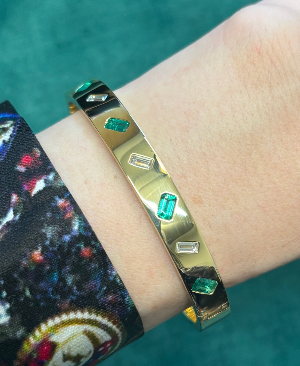 The Alternating Emerald Cut Diamond and Emerald Bangle in Yellow Gold adorns a wrist against a green background, complemented by a dark, patterned sleeve.