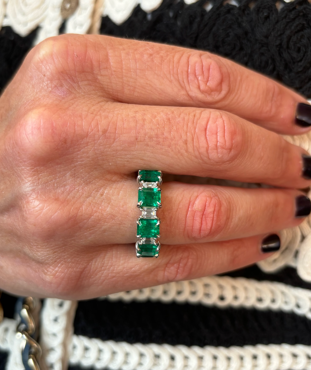 A hand with dark nail polish wears the Alternating Emerald Cut Diamond and Emerald Eternity Band in 18k white gold. The background is a black and white crocheted or knitted fabric.