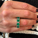 A hand with dark nail polish wears the Alternating Emerald Cut Diamond and Emerald Eternity Band in 18k white gold. The background is a black and white crocheted or knitted fabric.