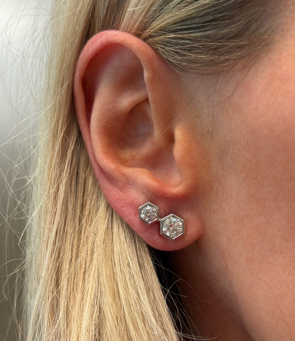 A close-up of a woman's ear adorned with Hexagon Bezel Set Round Diamond Duo Studs in 18k gold. Two duo studs are worn in each lobe, with her blonde hair softly framing her face and partially covering one side.