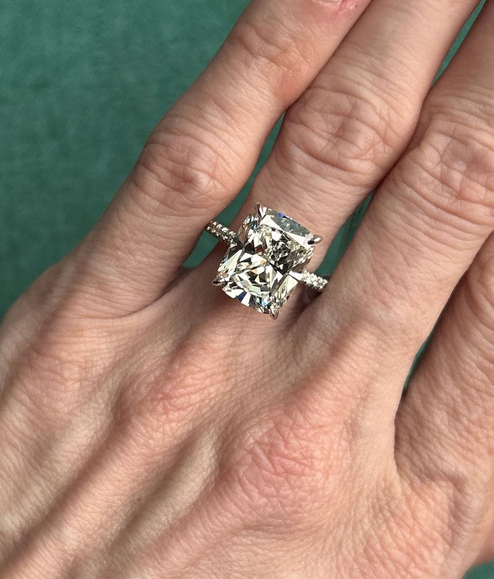 A hand wearing a Cushion Cut Diamond Engagement Ring with Pavé Band, displayed against a green background.
