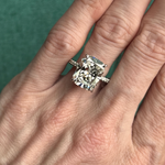 A hand wearing a Cushion Cut Diamond Engagement Ring with Pavé Band, displayed against a green background.