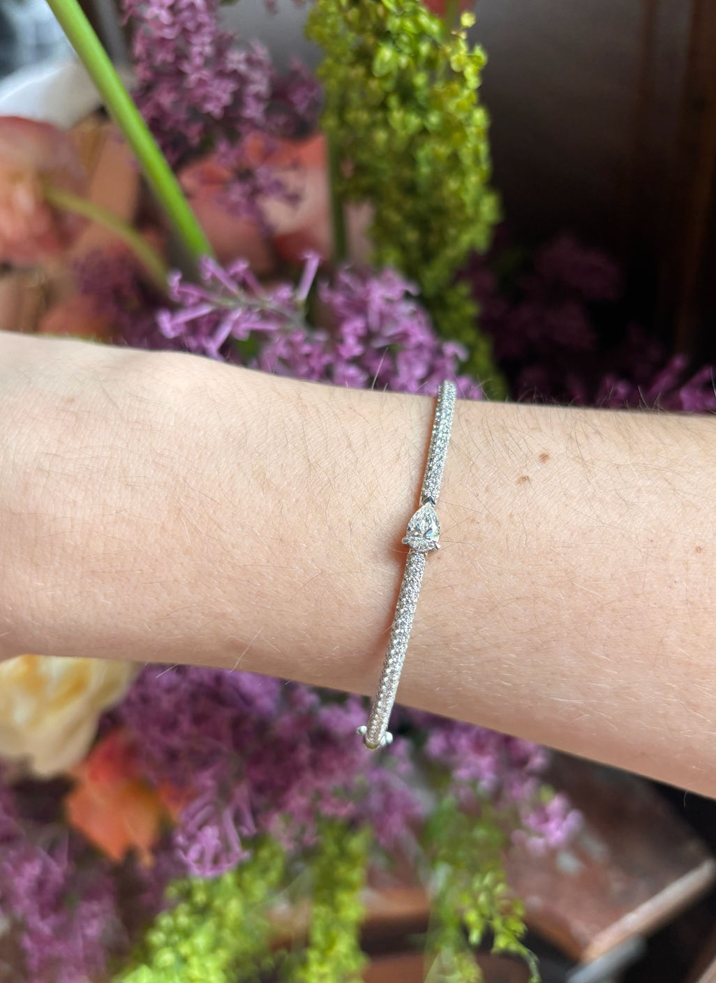 The East-West Set Pear Shape Diamond Pavé Bangle, featuring a teardrop diamond centerpiece, is elegantly worn on a wrist with blurred pink, purple, and green flowers in the background.