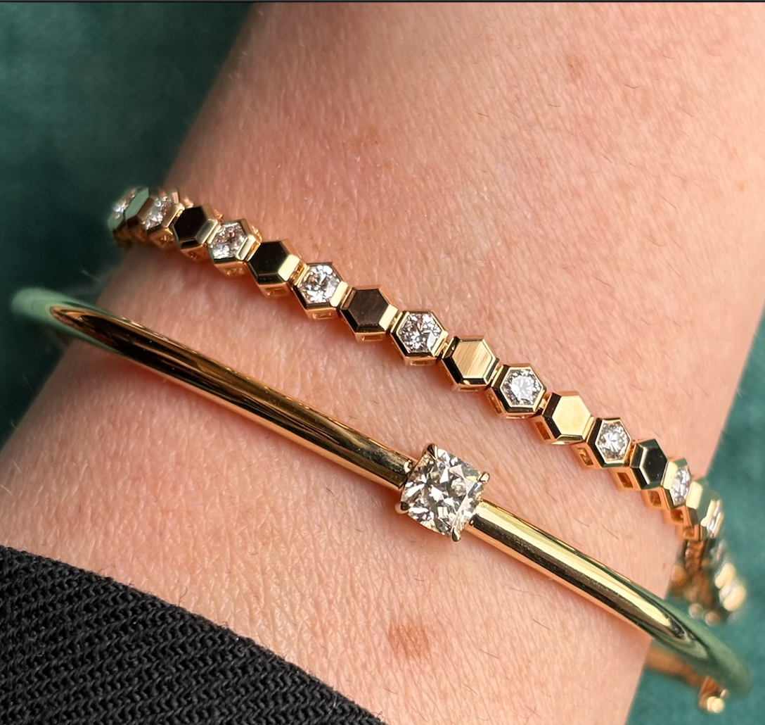 A close-up of a wrist wearing two gold bracelets: a Hexagon Bezel Set Bracelet with hexagon shape diamonds in yellow gold, and a simple gold bangle featuring a single center diamond.
