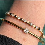 A close-up of a wrist wearing two gold bracelets: a Hexagon Bezel Set Bracelet with hexagon shape diamonds in yellow gold, and a simple gold bangle featuring a single center diamond.