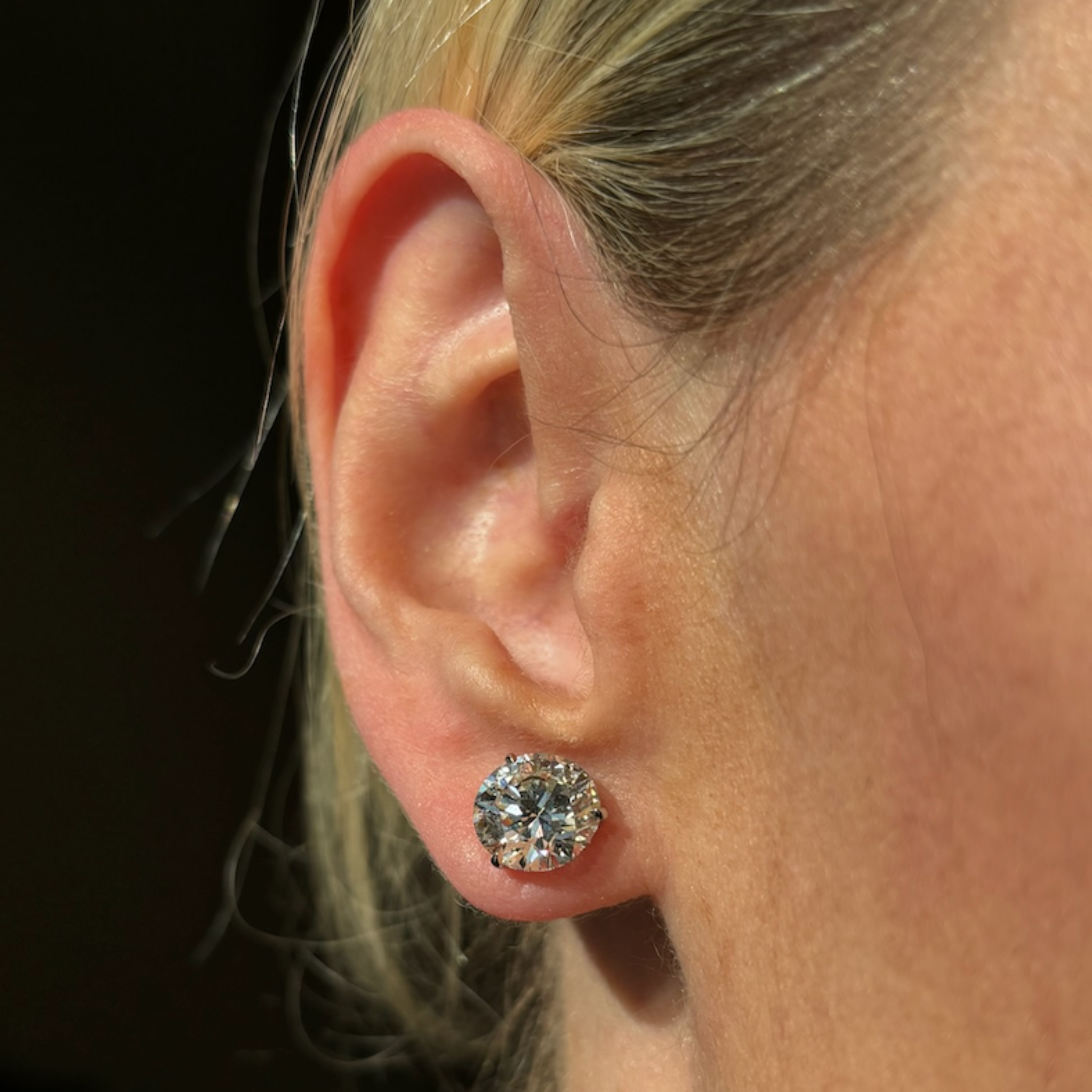 A close-up of a person with blonde hair wearing Classic Round Diamond Studs in White Gold. The large, brilliant diamonds are set in a sparkling martini setting, and the well-lit image highlights their radiant shine.