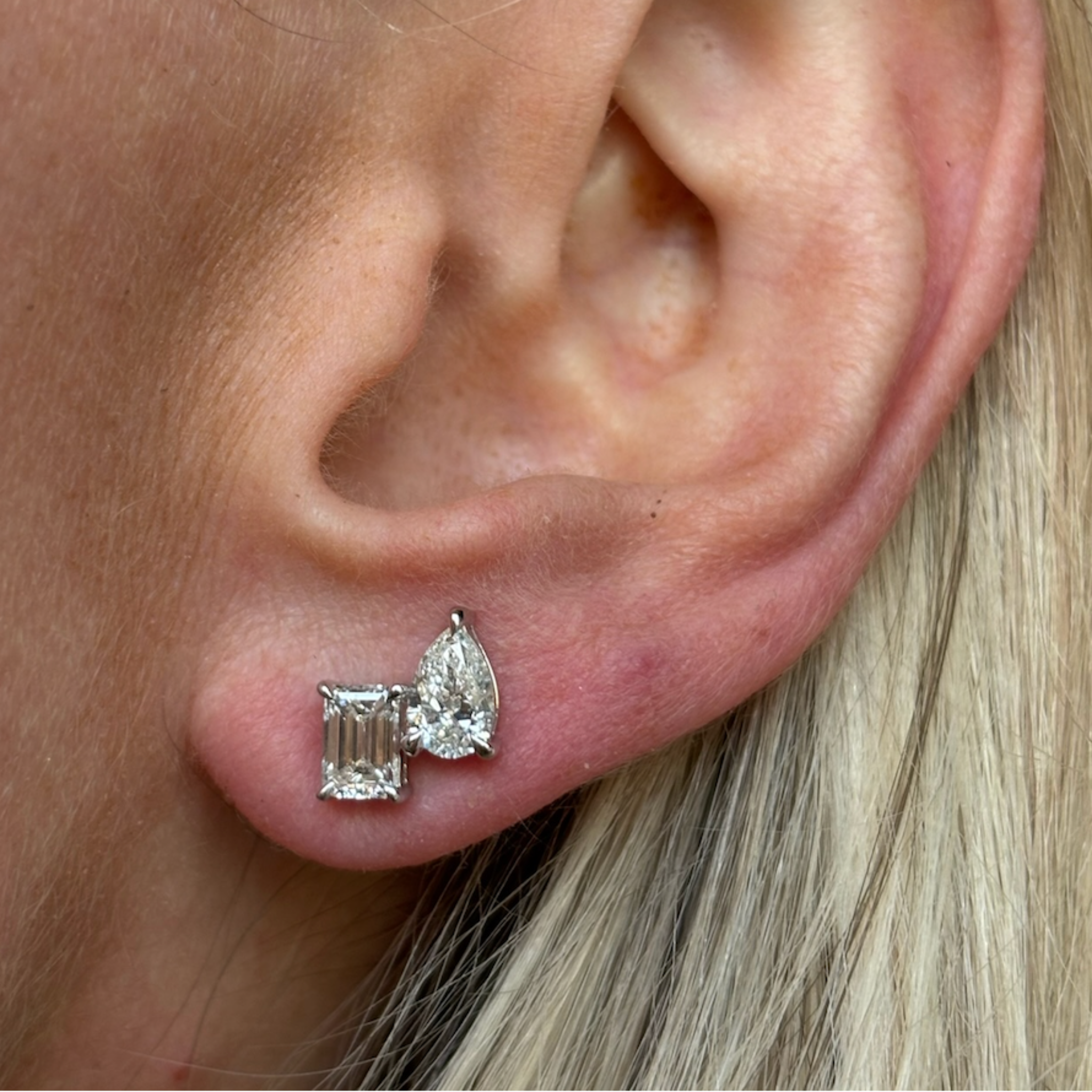 Close-up of a person's ear with blonde hair and light skin, wearing Mixed Shape Duo Studs featuring emerald cut and pear shape diamonds in white gold.