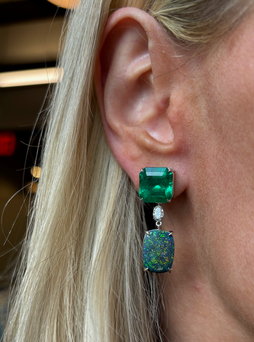Close-up of a woman's ear wearing Asscher Cut Sapphire, Emerald, Oval Boulder Opal, and Diamond Drop Earrings in White Gold. Blonde hair partially visible; earrings feature gemstones with iridescent and sparkling details.