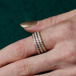 A close-up of a hand with gold-painted nails wearing three stacked rings, featuring the Hexagon Bezel Set Diamond Band encrusted with diamonds, against a dark green textured background.