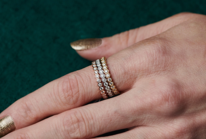 A close-up of a hand with gold-painted nails wearing three stacked rings, featuring the Hexagon Bezel Set Diamond Band encrusted with diamonds, against a dark green textured background.
