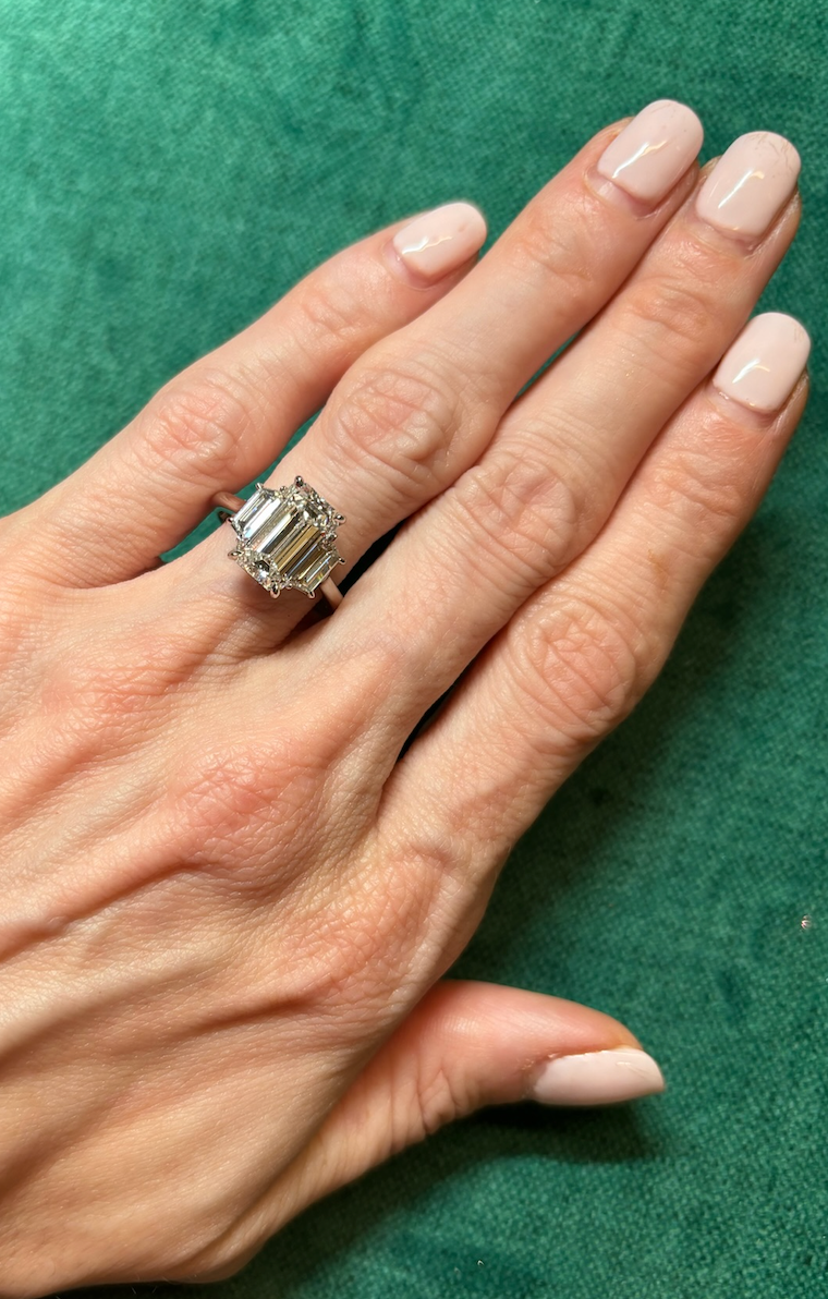 A hand with light skin and pale pink nails rests on green fabric, wearing the Emerald Cut Diamond Engagement Ring with Trapezoid Side Stones on the ring finger.