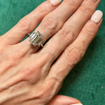 A hand with light skin and pale pink nails rests on green fabric, wearing the Emerald Cut Diamond Engagement Ring with Trapezoid Side Stones on the ring finger.