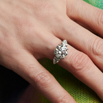 A hand with gold glitter nail polish showcases the Round Brilliant Diamond Engagement Ring with Heart Side Stones on the ring finger, set against a vibrant green and pink background.