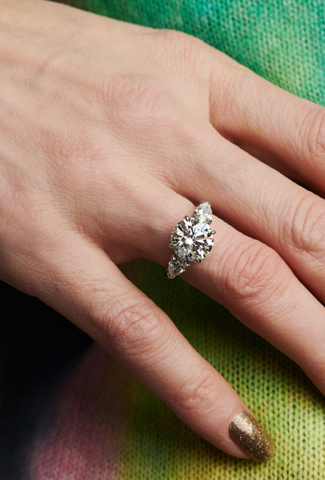 A hand with gold glitter nail polish showcases the Round Brilliant Diamond Engagement Ring with Heart Side Stones on the ring finger, set against a vibrant green and pink background.