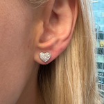 A close-up of a person’s ear wearing Bezel Set Diamond Heart Studs in 18k gold. Their straight, blonde hair frames the sparkling earrings, with a softly blurred glass building visible in the background.