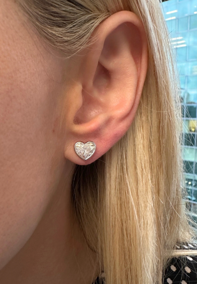 A close-up shows a person with straight blonde hair wearing Bezel Set Diamond Heart Studs in White Gold, with a building's glass windows visible in the background.