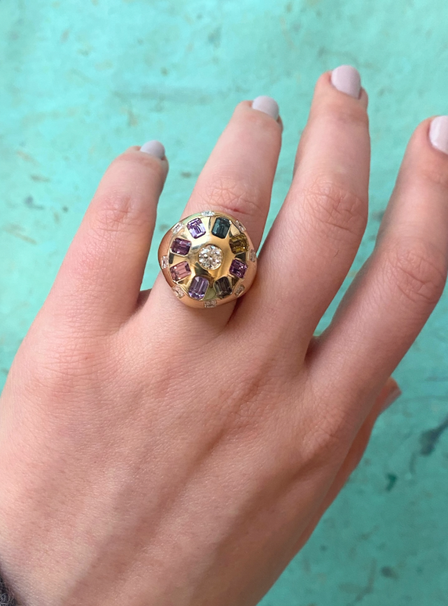 A hand with pale nail polish wears the Confetti Bombé Ring in 18k yellow gold, featuring a large central diamond and colorful gemstones, set against a turquoise background.
