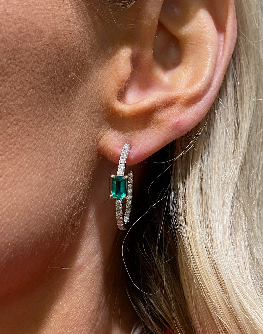Close-up of a person's ear with blonde hair wearing Pavé Hoops with Emerald Cut Emeralds in White Gold, featuring diamond pavé detailing and a striking rectangular green gemstone at the center.