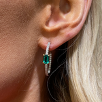 Close-up of a person's ear with blonde hair wearing Pavé Hoops with Emerald Cut Emeralds in White Gold, featuring diamond pavé detailing and a striking rectangular green gemstone at the center.