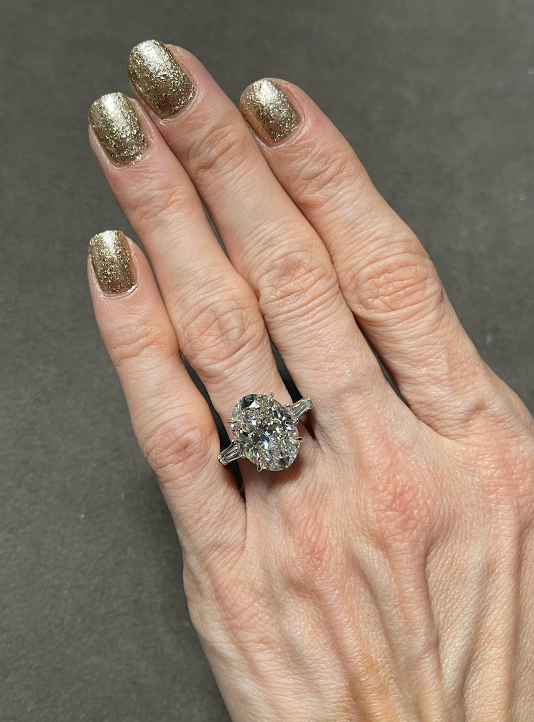 A hand with gold glitter nails displays the Oval Shape Diamond Engagement Ring with Tapered Baguette Side Stones, set in platinum, against a dark background.
