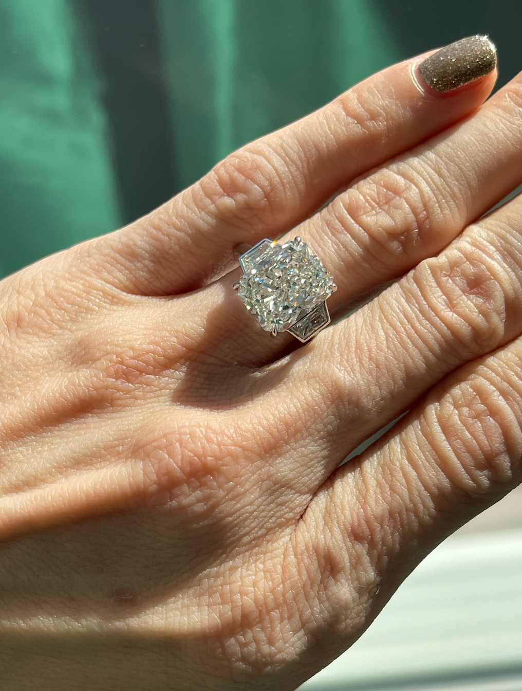 A hand with manicured nails wears the Radiant Cut Diamond Engagement Ring with Bezel Set Epaulette Side Stones, its silver band sparkling in natural sunlight.