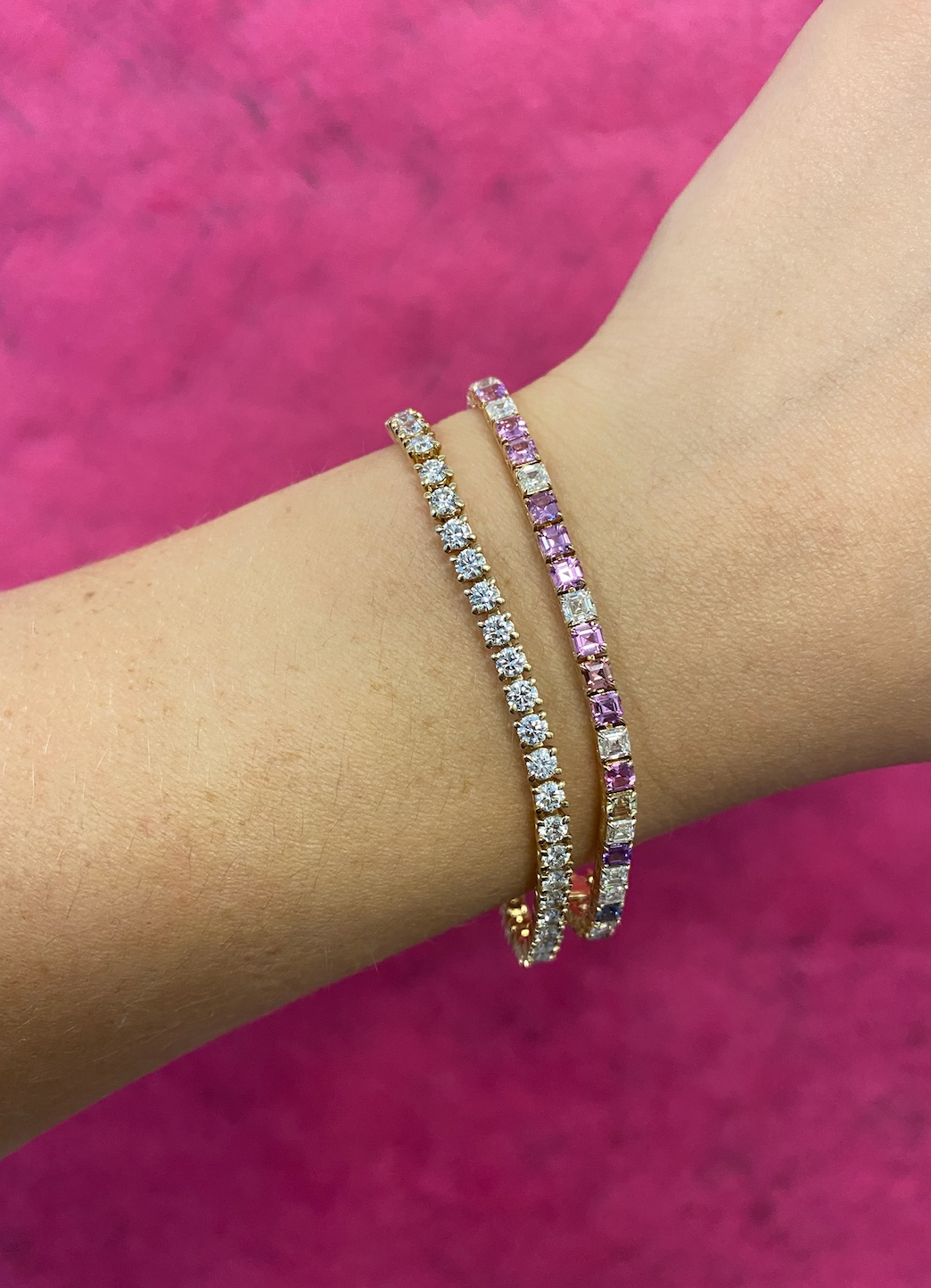 A close-up of a wrist wearing two tennis bracelets, including the Multicolor Asscher Cut Diamond and Sapphire Tennis Bracelet in Yellow Gold, set against a bright pink background.