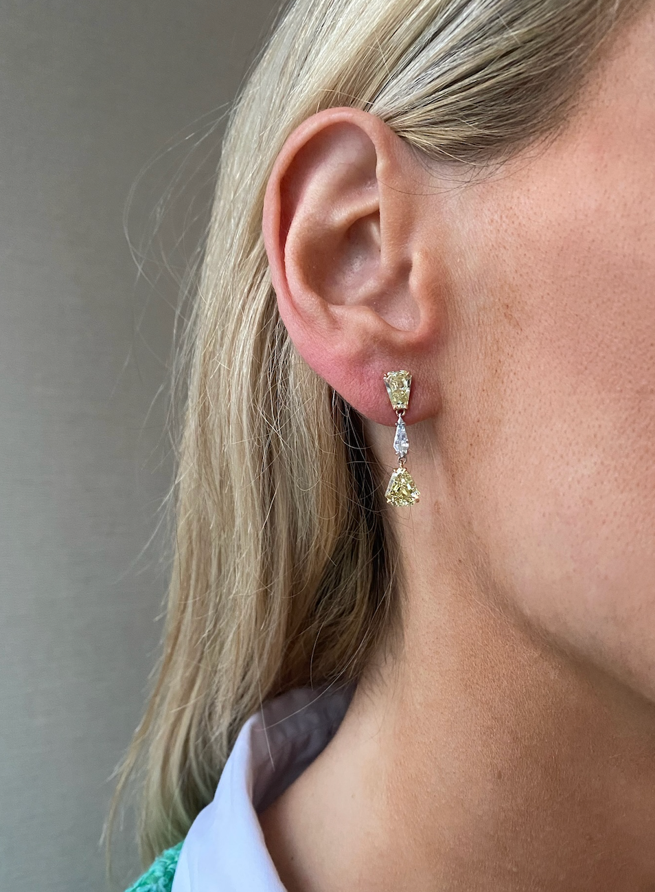 Close-up of a woman's ear wearing This & That Champagne Flute Drop Earrings with fancy yellow and white mixed shape diamonds, set in 18K yellow gold. She has blonde hair and wears a white collared shirt.