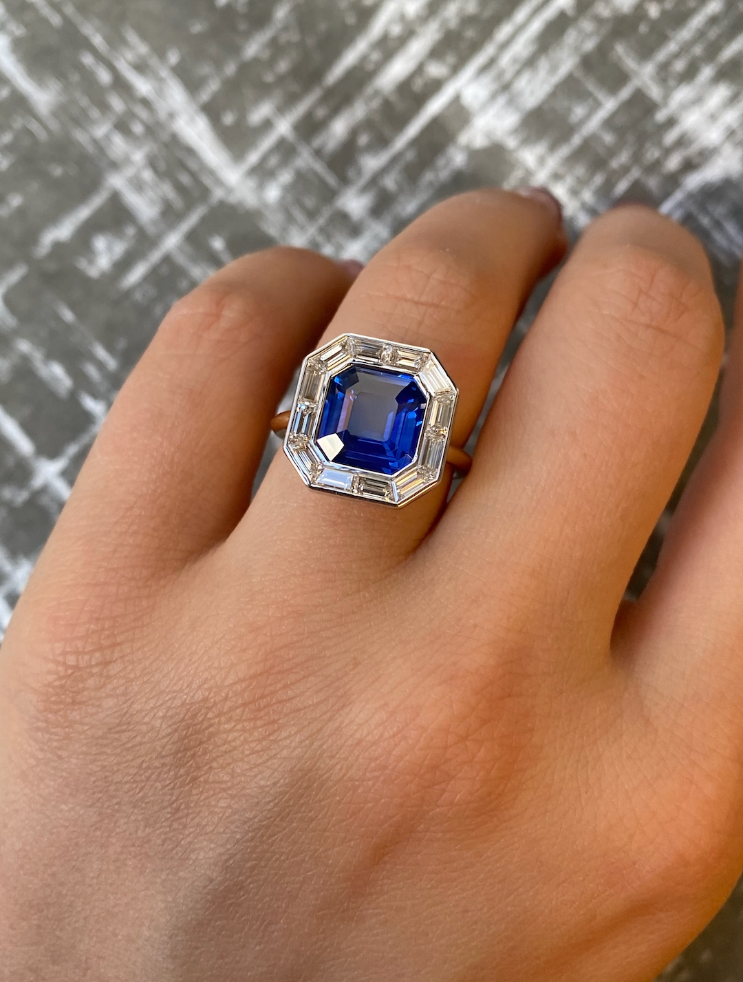 A hand wears the Mosaic Ring with an Asscher cut blue sapphire and baguette diamonds, set in 18K gold, against a gray textured background.