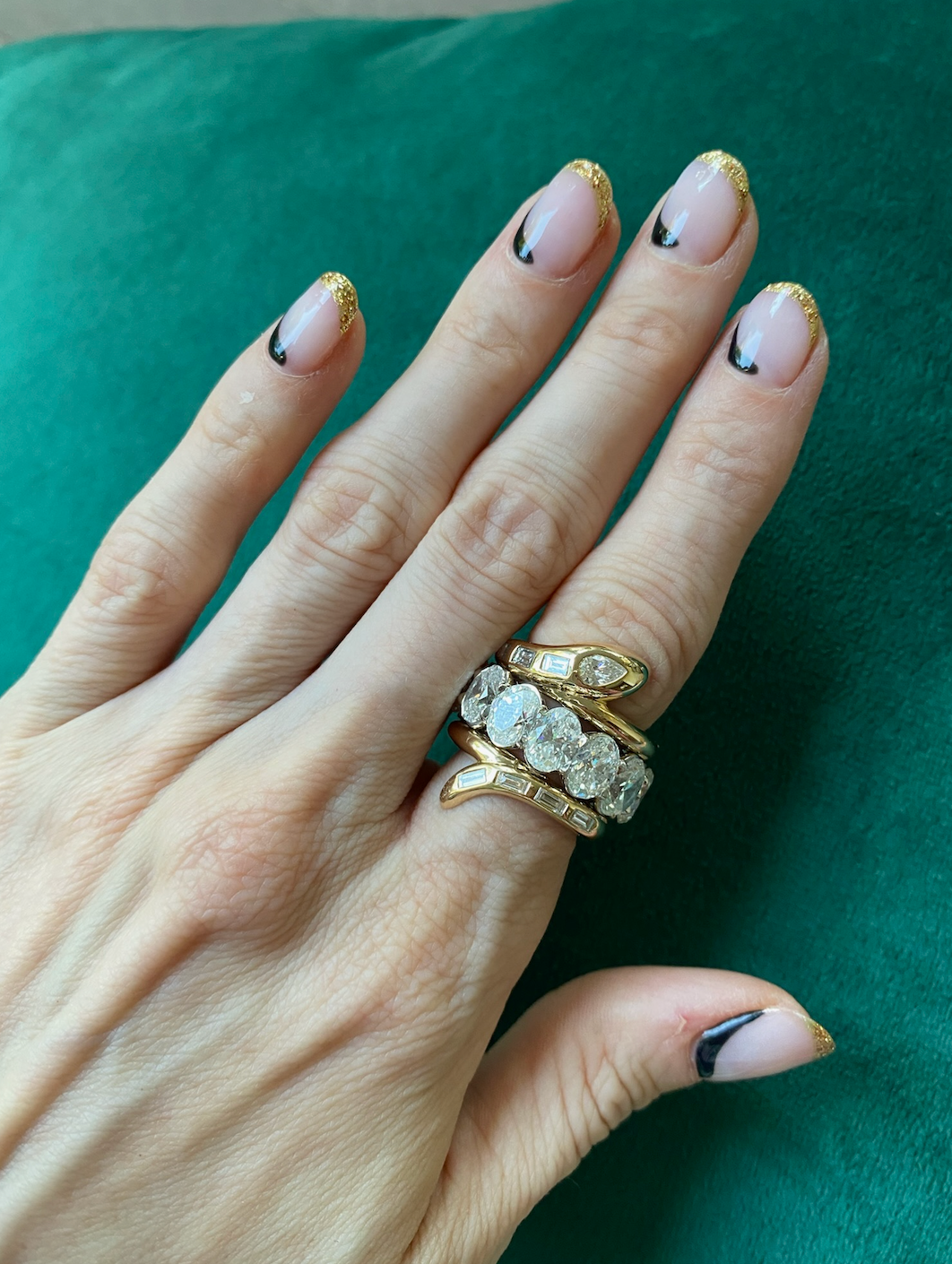 A hand wearing Serpent Bookend Rings in yellow gold with mixed shape diamonds rests on green. Nails feature gold glitter French tips and a curved black detail near the cuticle.