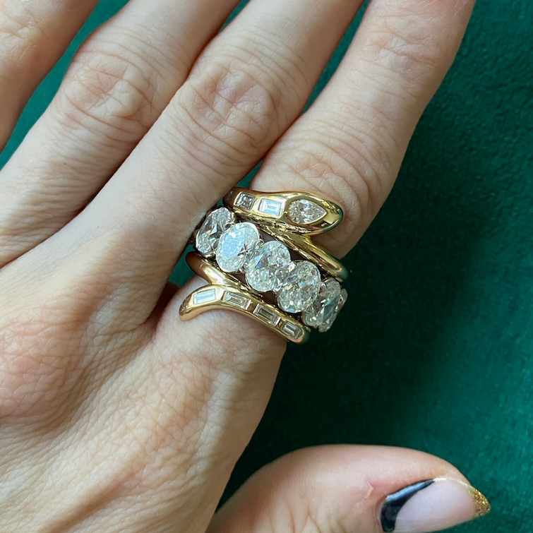 A hand wearing Serpent Bookend Rings in yellow gold with mixed shape diamonds rests on green. Nails feature gold glitter French tips and a curved black detail near the cuticle.