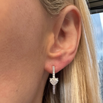 Close-up of a woman's ear with straight blonde hair, wearing the Heart Diamond Pavé Hoops—18K white gold pavé hoops with a large heart-shaped diamond center. City buildings and a window appear blurred in the background.