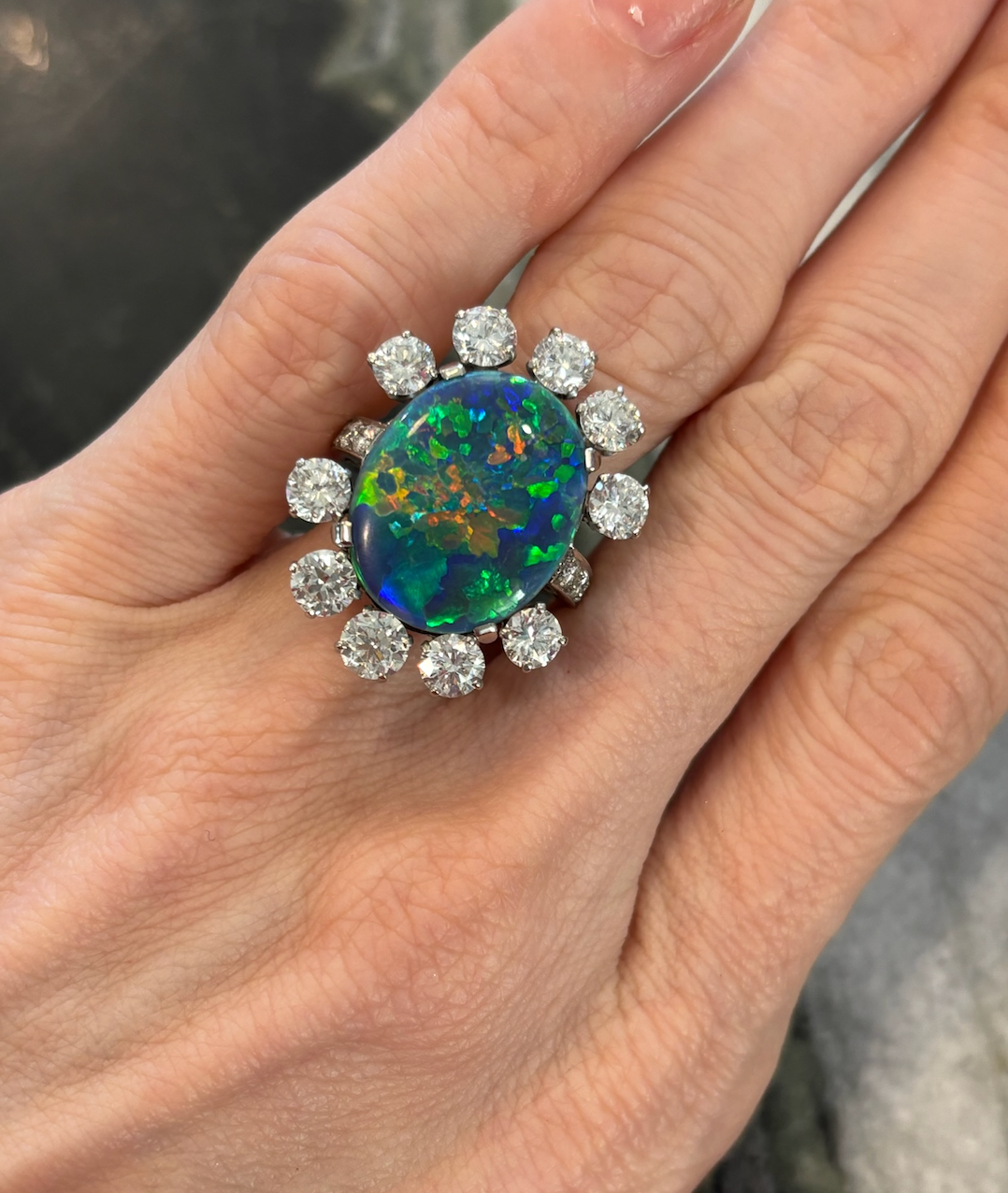 A hand wearing the Sunburst Oval Opal Ring with vivid blue, green, and red hues, encircled by round brilliant diamonds and pavé, set in 18K white gold.