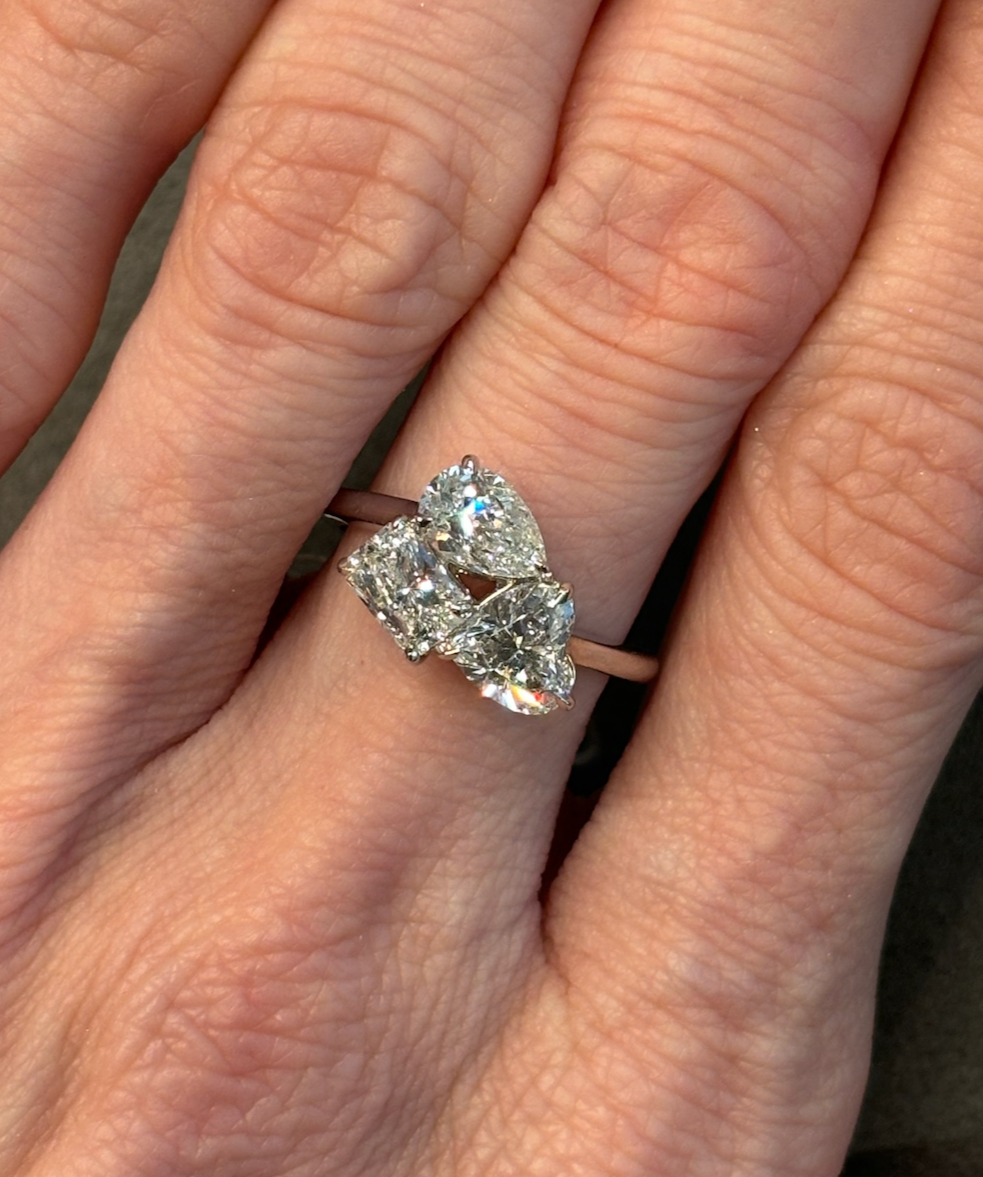 A hand wearing a Three Stone Ring in white gold featuring pear-shaped, heart-shaped, and radiant cut diamonds on the ring finger.