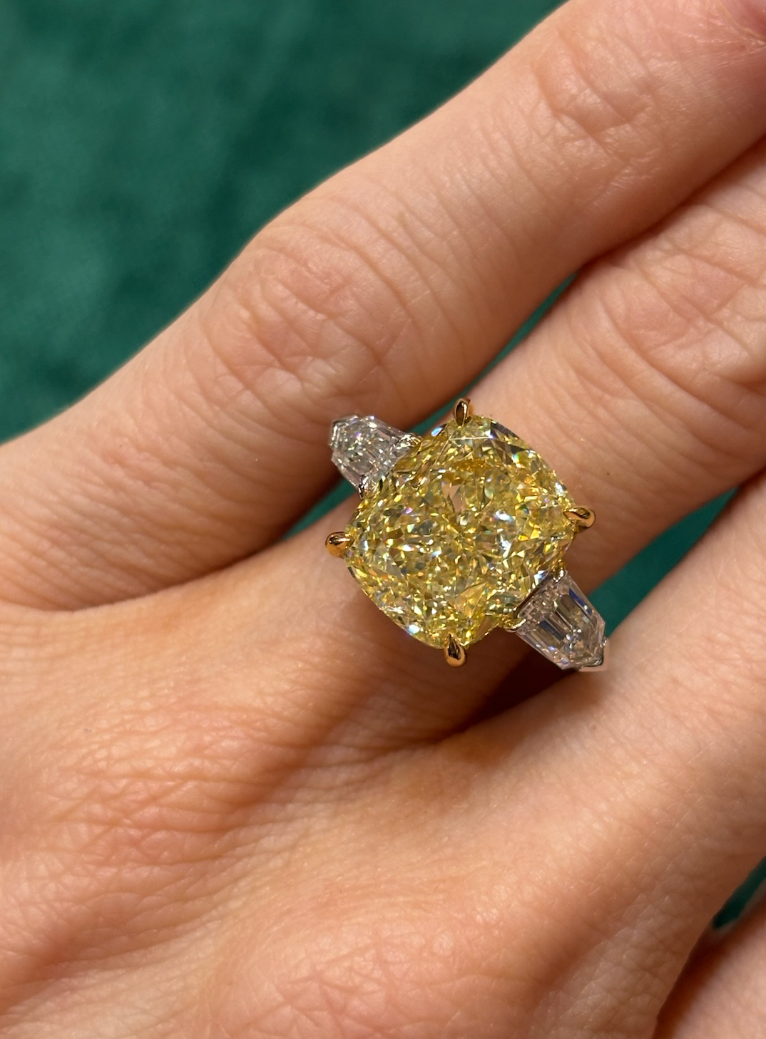 A hand displays the Fancy Yellow Radiant Cut Diamond Engagement Ring with tapered baguette side stones, featuring a vibrant yellow center stone against a green background.