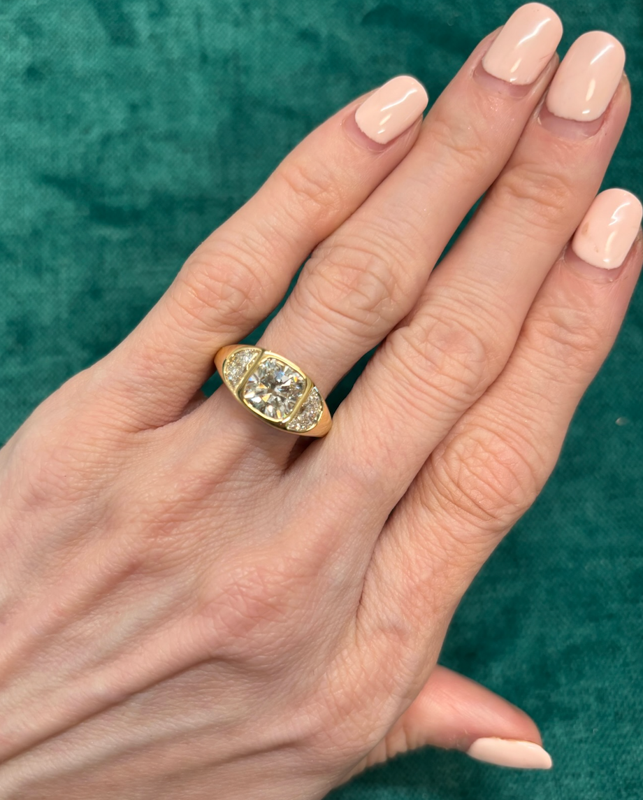 A hand with light pink polished nails wears the Burnish Set Cushion Diamond Engagement Ring with Half Moon Side Stones, set against a textured green background.