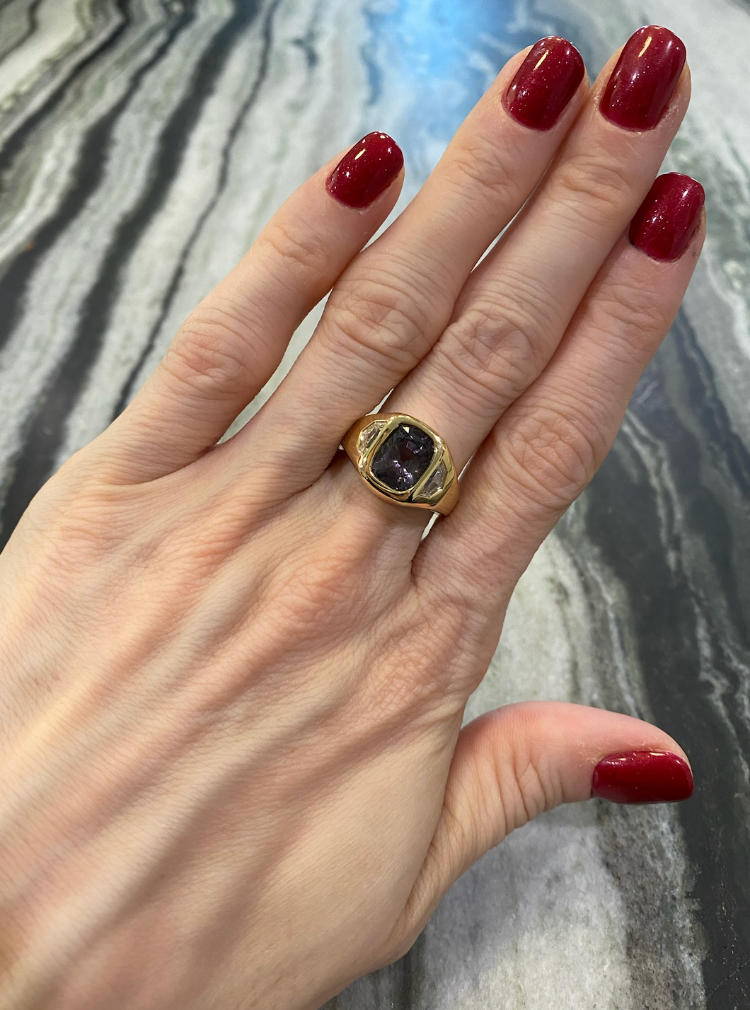 A hand with red painted nails wears the Gypsy Ring in yellow gold, featuring a cushion cut spinel and diamond epaulette side stones, set against a gray and white marbled background.