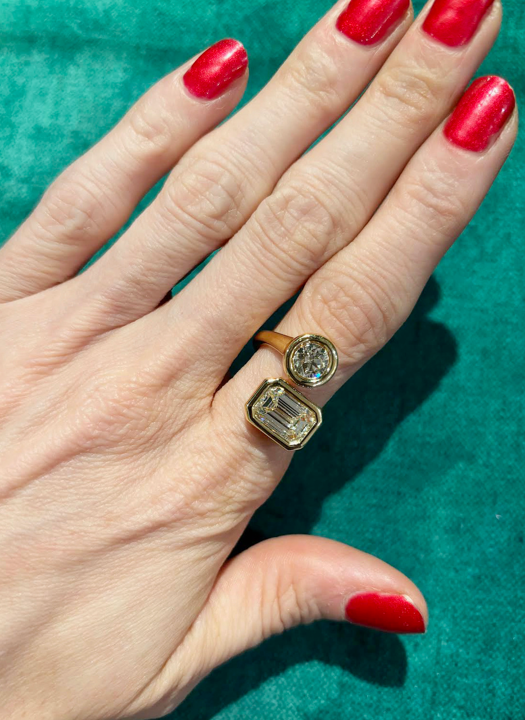 A hand with bright red nail polish wears the Toi Et Moi Wraparound Ring Bezel Set in Yellow Gold with round and emerald cut diamonds against a teal background.