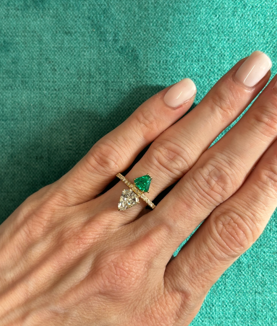 A hand with light pink nails wears the Emerald and Diamond Double Shield Ring in Yellow Gold, featuring pear-shaped green and clear gemstones separated by a horizontal bar, set against a teal fabric background.