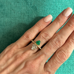 A hand with light pink nails wears the Emerald and Diamond Double Shield Ring in Yellow Gold, featuring pear-shaped green and clear gemstones separated by a horizontal bar, set against a teal fabric background.
