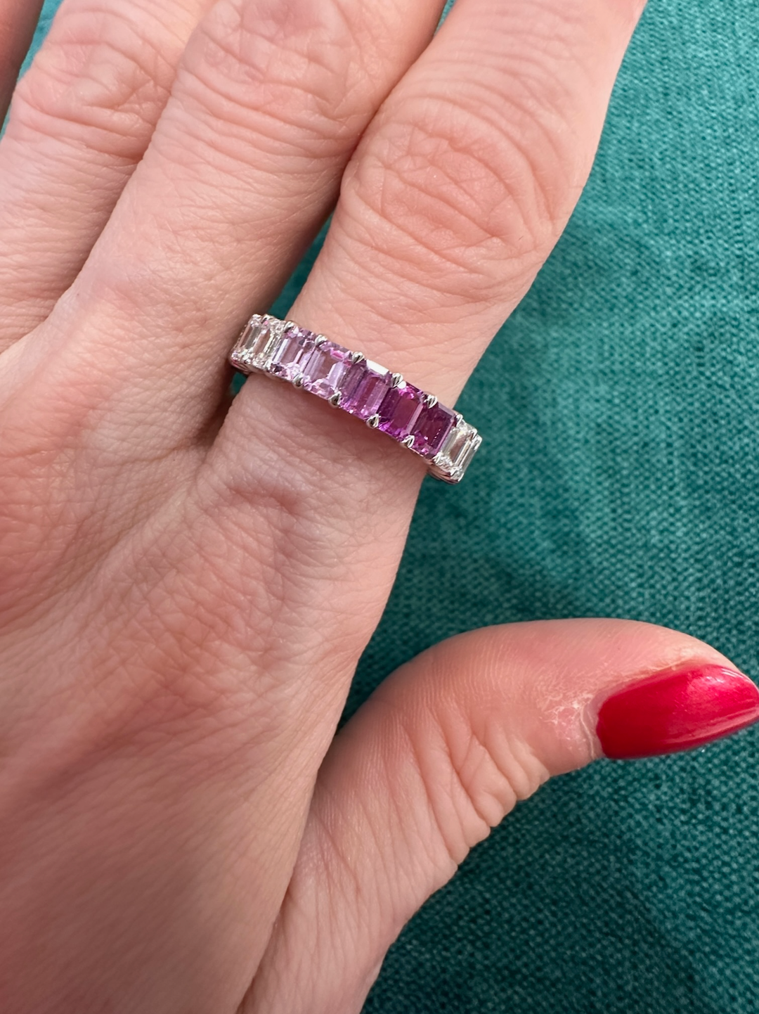 A hand with red-painted nails wears the Ombré Eternity Band with Emerald Cut Pink Sapphires and White Diamonds, shown against a green fabric background.