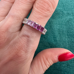 A hand with red-painted nails wears the Ombré Eternity Band with Emerald Cut Pink Sapphires and White Diamonds, shown against a green fabric background.