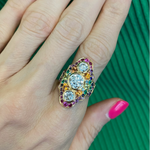 A hand with pink nails showcases the Multicolor Dinner Ring in Yellow Gold, featuring sapphires and three round diamonds, against a green ribbed fabric background.