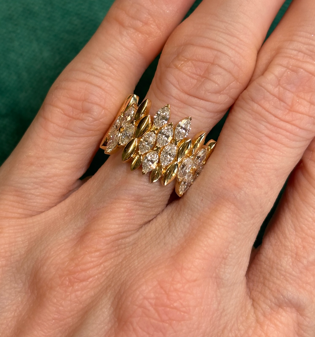 A close-up of a hand wearing the Multi Tier Cluster Marquise Eternity Band in yellow gold, featuring marquise-shaped accents and diamond clusters, set against a green background.